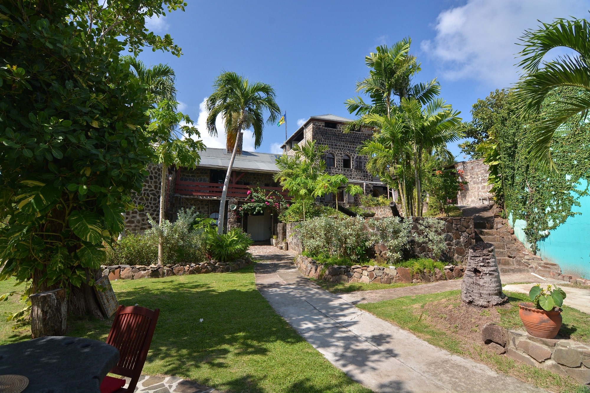 A stone building with a tin roof is surrounded by palm trees and lush greenery under a blue sky. This Caribbean Estate Leasehold Opportunity features a stone path through the well-kept garden with outdoor seating and vibrant tropical plants. Photo