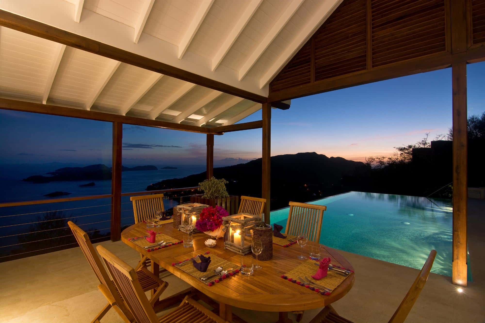 A wooden dining table set for dinner under a covered patio overlooks an infinity pool and a scenic sea view at sunset, with warm lighting creating a cosy ambience—perfect for guests enjoying a luxury villa hire in Bequia.