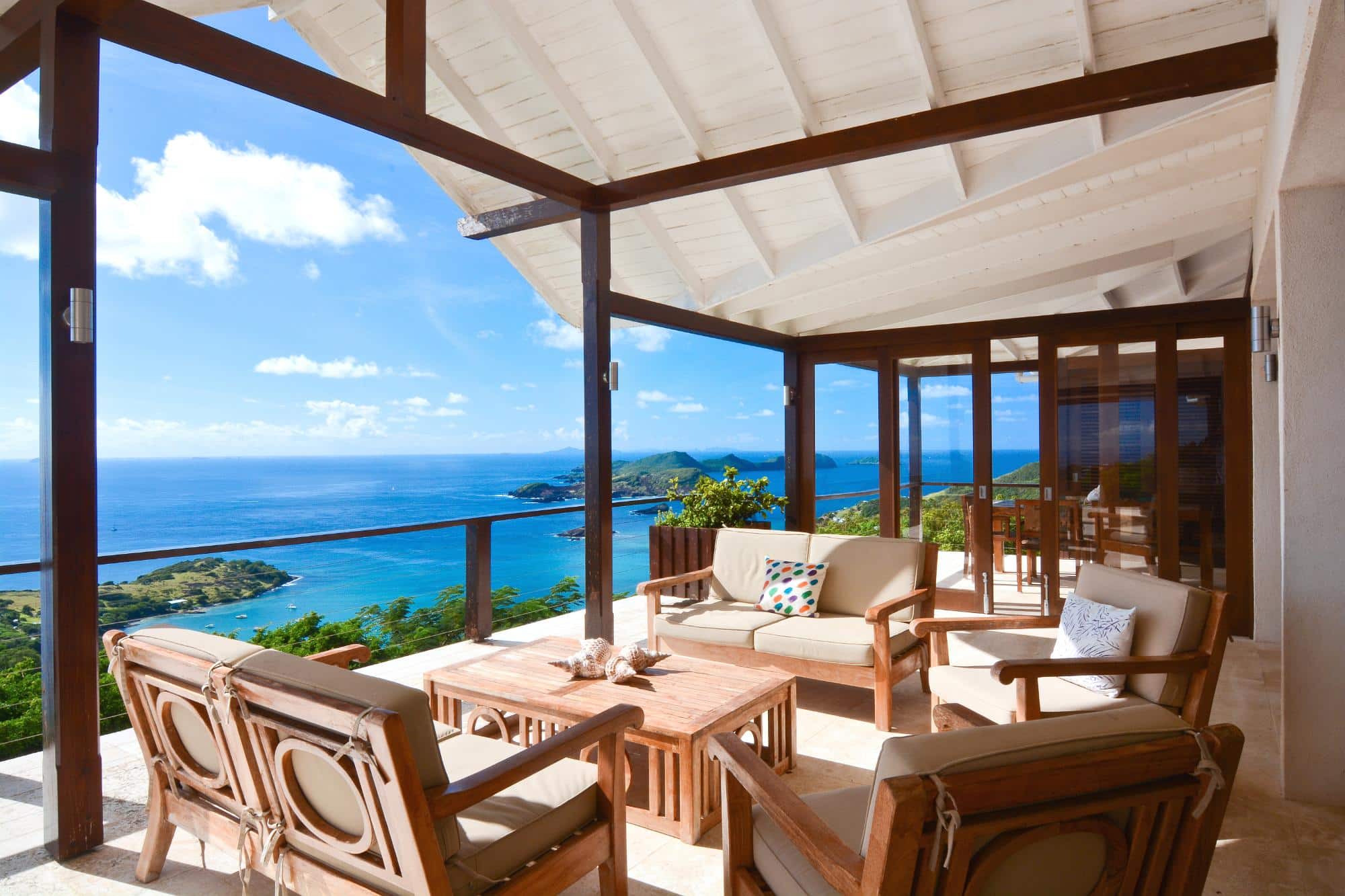 A covered patio with wooden furniture overlooks a blue sea, green hills, and a partly cloudy sky. The patio has cushioned chairs, a sofa, a wooden table, and glass doors leading inside.