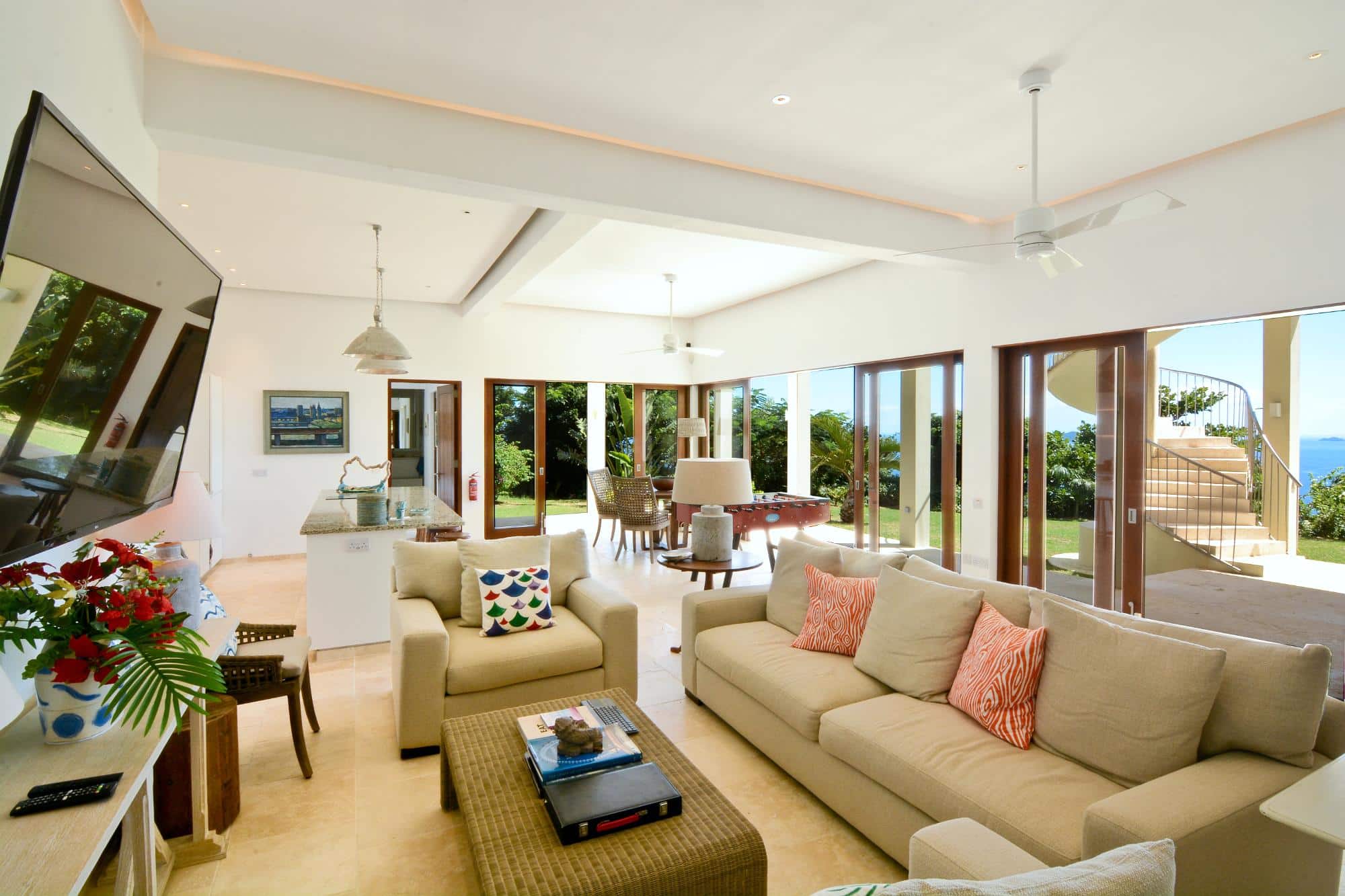 Bright, airy living room with beige sofas, colourful cushions, a television, and large windows overlooking a lush garden and the sea—perfect for your luxury villa rental Bequia escape. Dining area and kitchen are visible in the background.