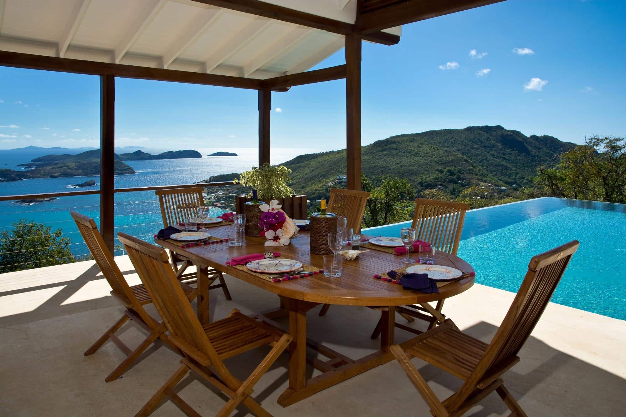 Outdoor dining area with a wooden table and chairs set beside an infinity pool, overlooking mountains, sea, and islands under a clear blue sky. Table is set with plates, glasses, and a vase of flowers.