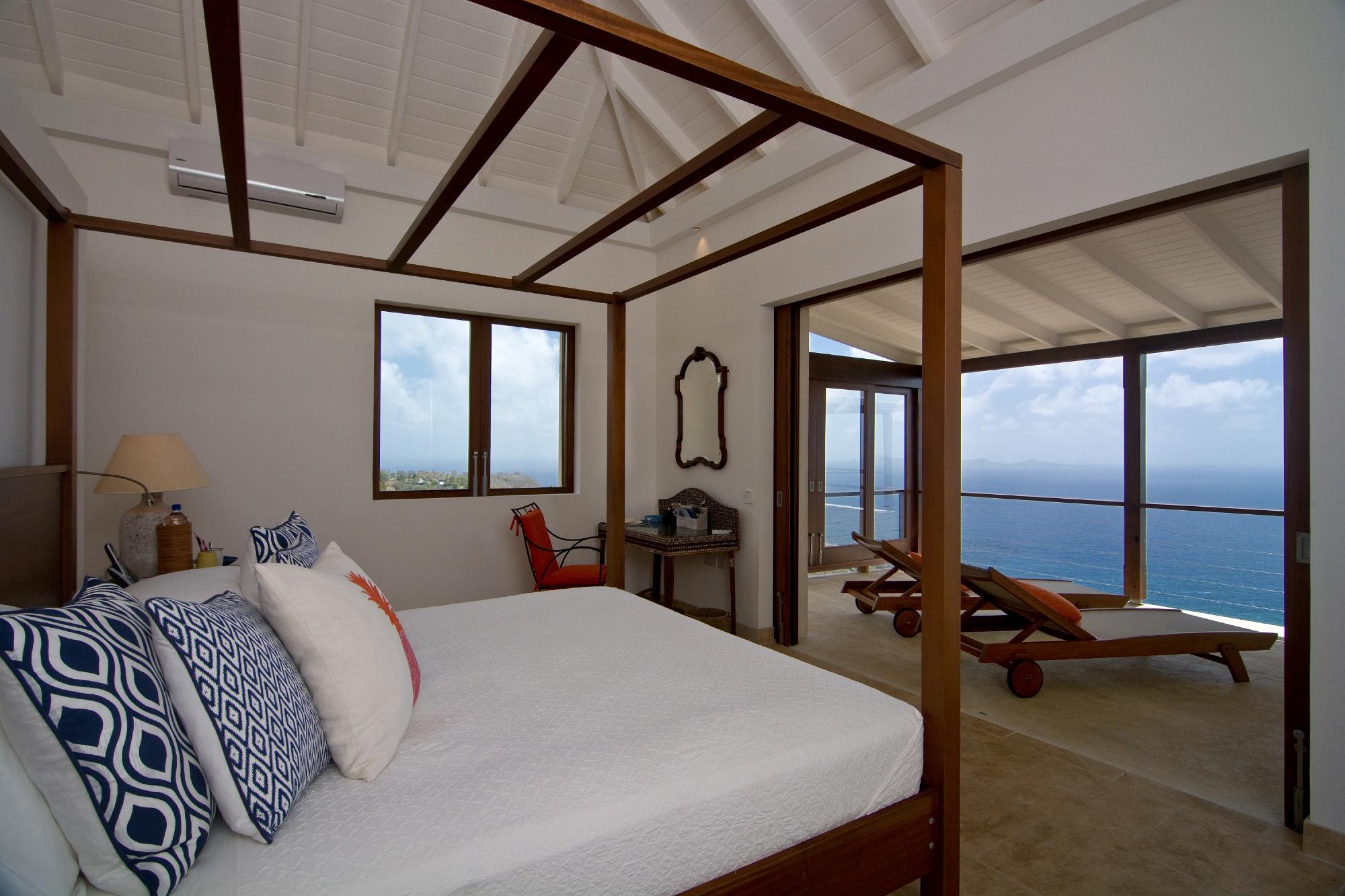 A bright bedroom in a luxury villa rental Bequia with a four-poster bed, patterned cushions, and sweeping sea view. Sliding doors open to a terrace with loungers overlooking the sea. White walls and high ceilings complete the airy look.