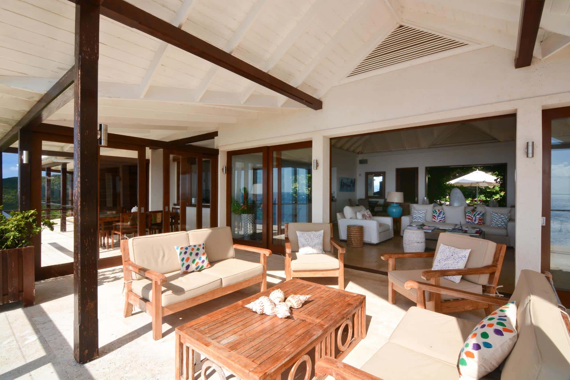 A sunlit patio with wooden furniture and cushions leads into a spacious, open-plan living room with sofas and elegant décor, all overlooking a scenic view through glass doors—perfect for those seeking a luxury villa hire in Bequia.