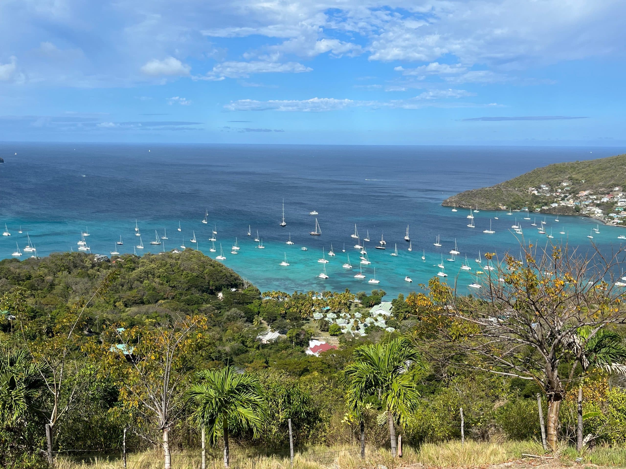 Many sailboats anchored in a turquoise bay near a villa for sale in Mount Pleasant, Bequia, surrounded by hills covered in greenery.
